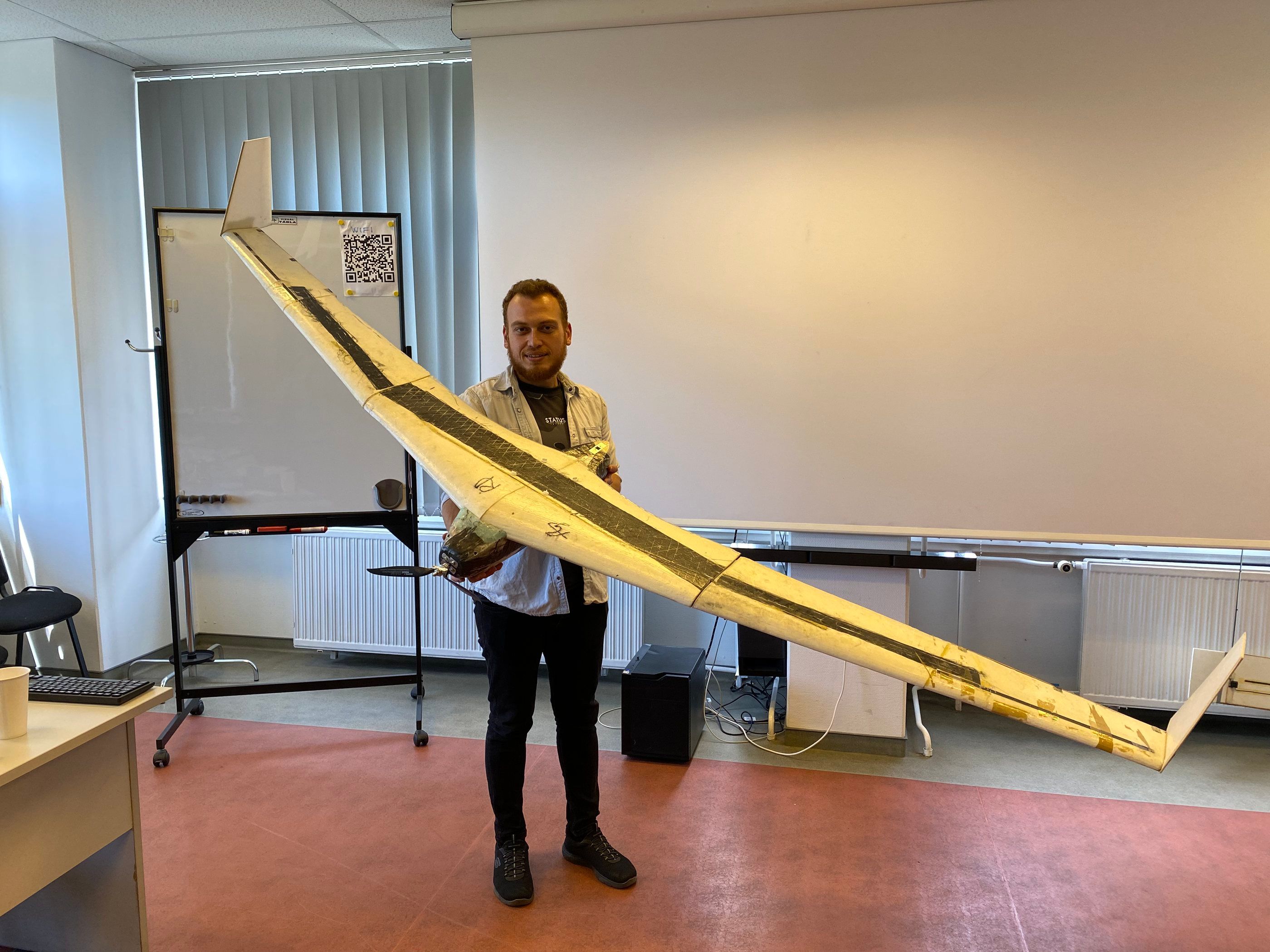 Emre Özbek holding a large flying-wing glider in a classroom